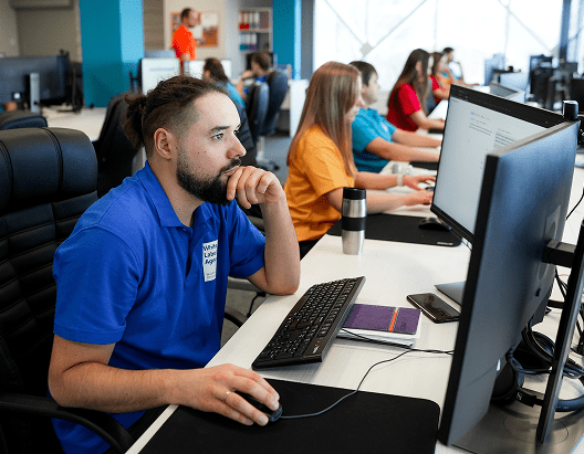 A man with a beard and hair bun wearing a blue polo shirt, focused on his computer screen in a shared office space.