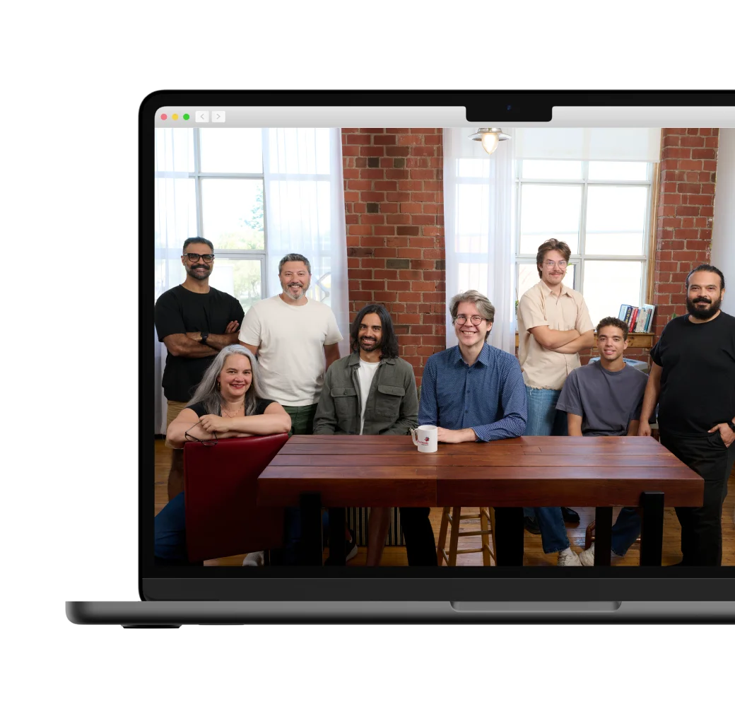 A group of eight people posing for a team photo in a bright office with brick walls and a large wooden table.