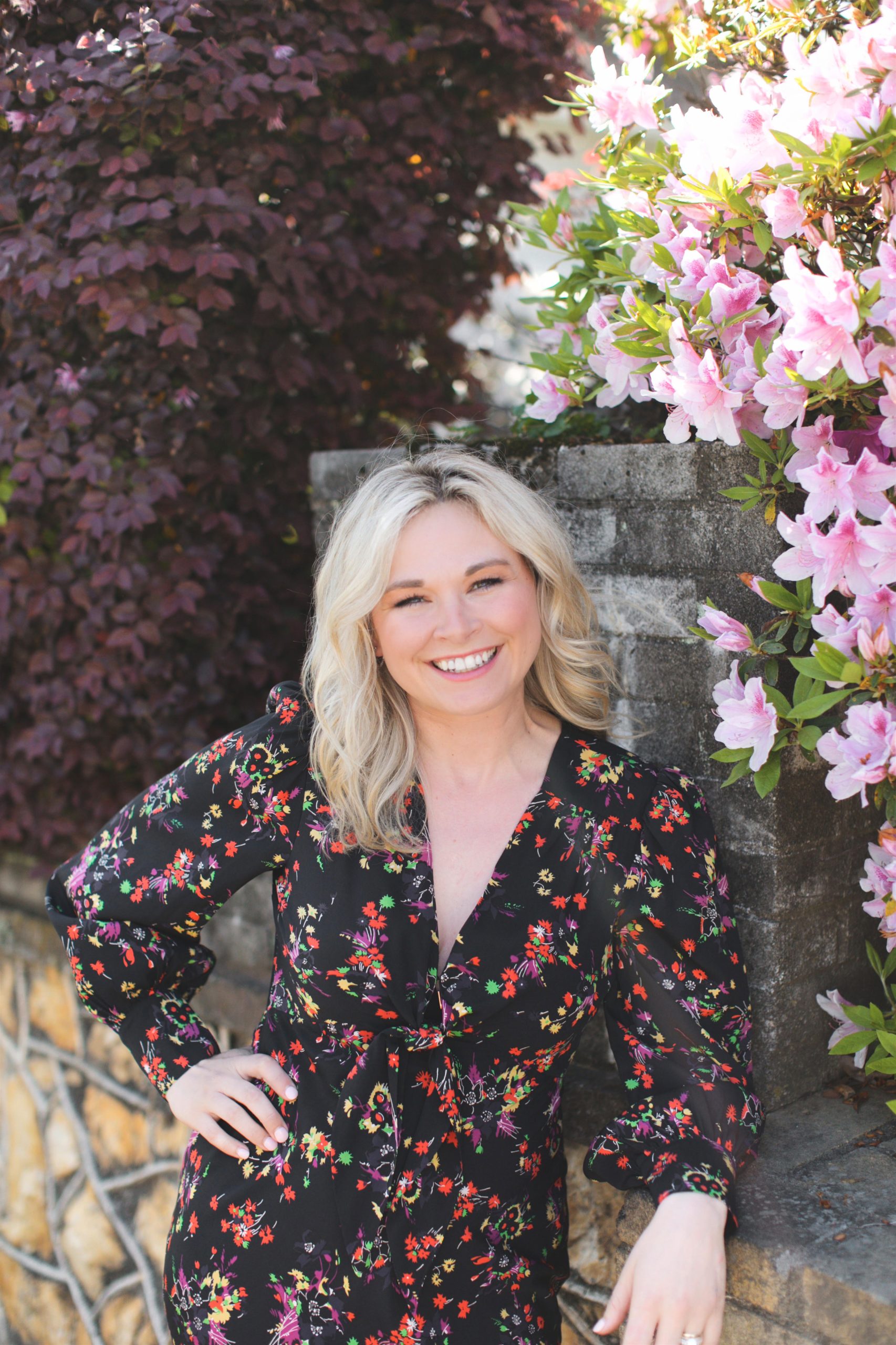 A smiling woman with blonde hair wearing a floral black dress, leaning against a stone wall surrounded by pink and purple flowers.