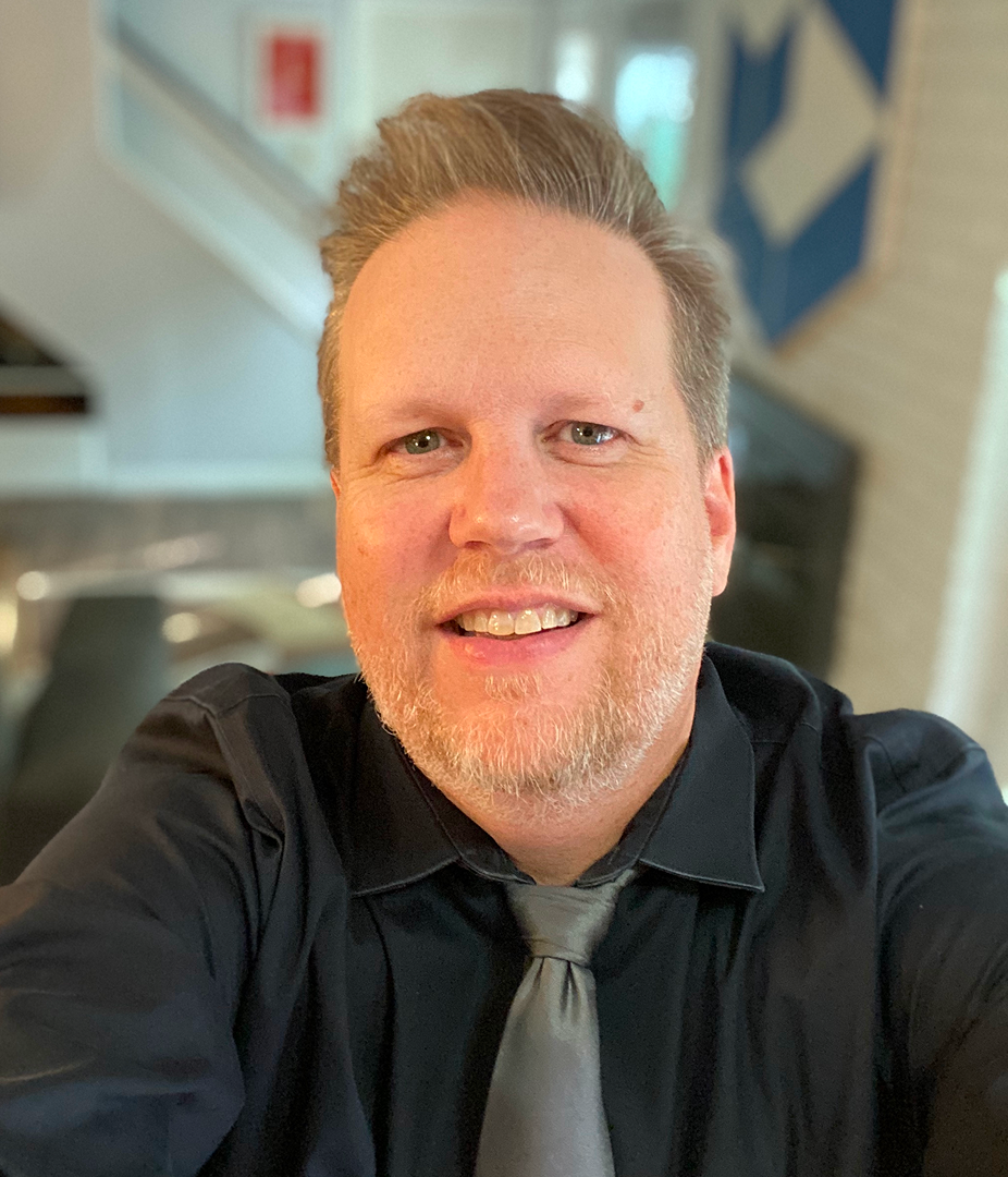 A close-up portrait of a smiling man with light hair and a blonde beard, wearing a black button-down shirt and a grey silk tie, in an indoor setting with a blurred background.