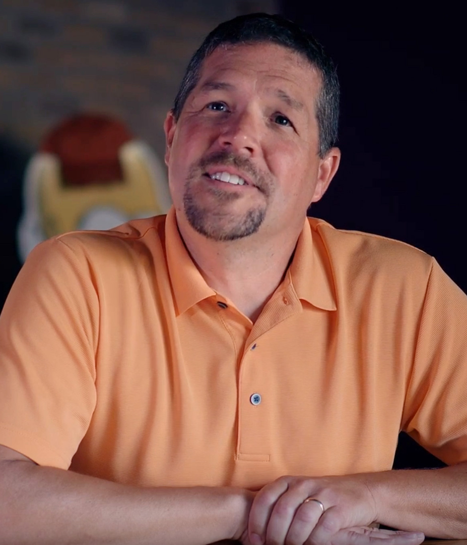 A smiling man with a goatee, wearing a light orange polo shirt, sitting with his hands clasped in front of him in an indoor setting with a dark, blurred background.